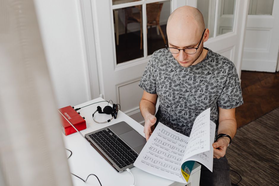 Adult male musician studying music sheets at a home desk with a laptop.