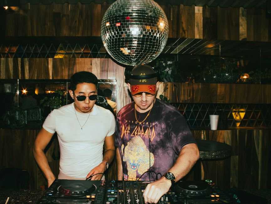 Two DJs mixing music at a vibrant club with a glittering disco ball overhead, creating an energetic nightlife scene.